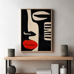 May include: Abstract art print featuring a black and white stylized face with a red lip. The print is framed and hanging on a white wall above a wooden shelf.