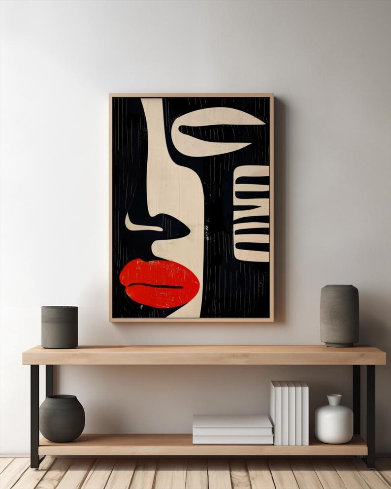 May include: Abstract art print featuring a black and white stylized face with a red lip. The print is framed and hanging on a white wall above a wooden shelf.
