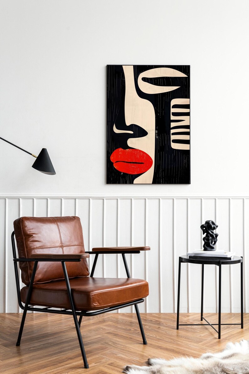 May include: A black and white abstract art print of a face with a red lip. The print is hanging on a white wall above a brown leather armchair and a black side table.
