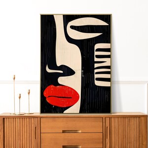 May include: Abstract art print of a face with red lips, black hair, and white and black geometric shapes. The print is framed in a gold frame and is hanging on a white wall.