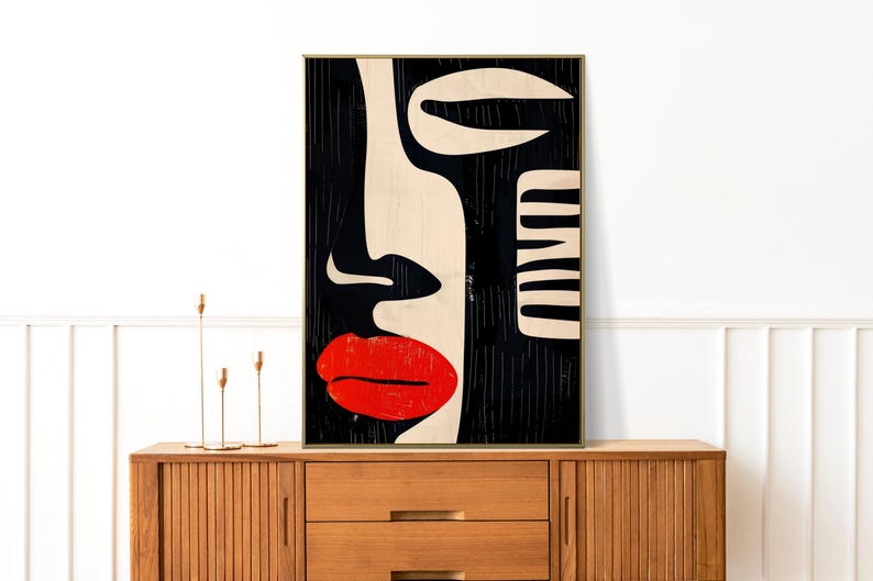 May include: Abstract art print of a face with red lips, black hair, and white and black geometric shapes. The print is framed in a gold frame and is hanging on a white wall.
