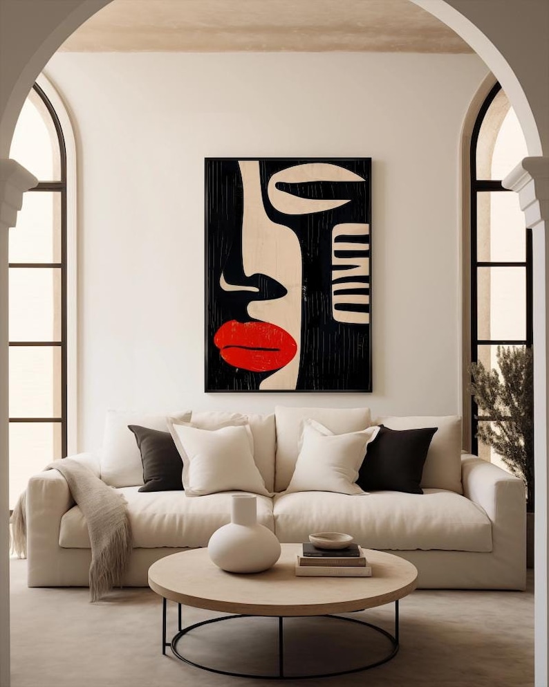 May include: A white couch with black and white pillows in a living room with a black and white abstract art print above it. The couch has a throw blanket on it and a round wooden coffee table with a vase and books on it.