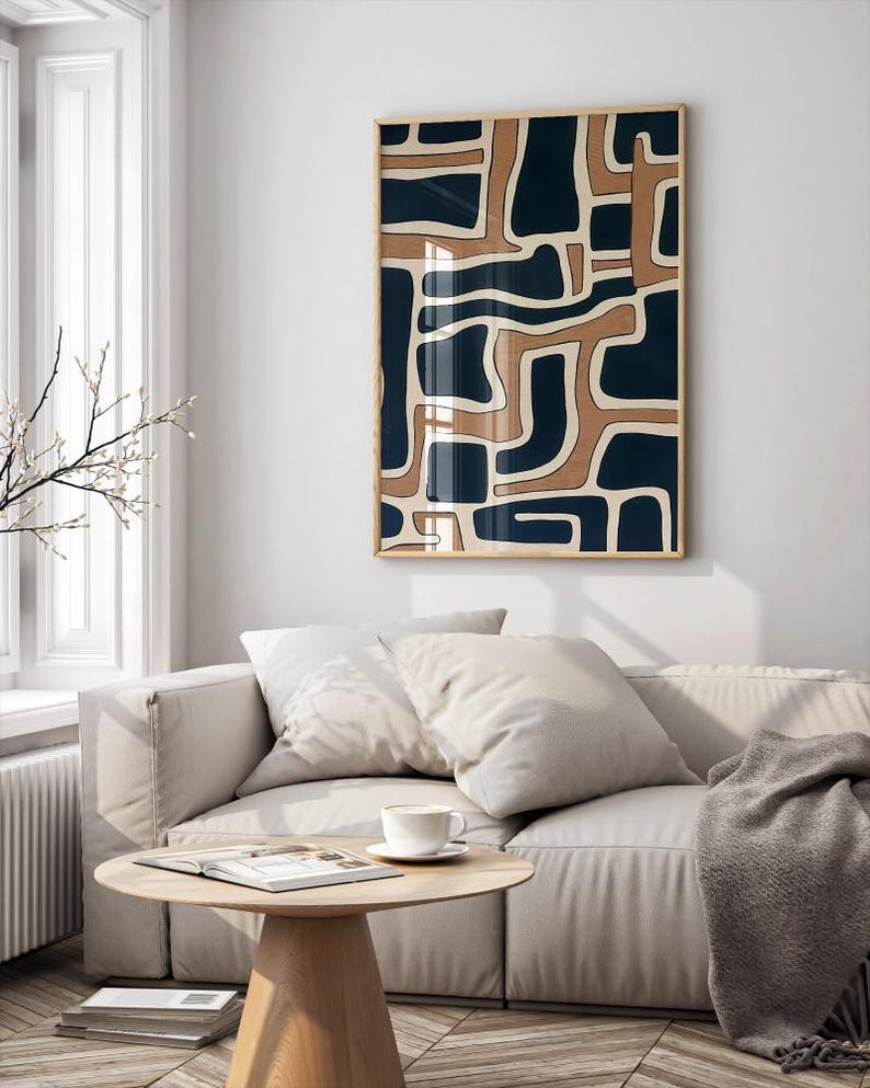 May include: A beige couch with two pillows and a gray throw blanket in front of a white wall with a framed abstract print featuring a geometric pattern of brown and blue shapes.