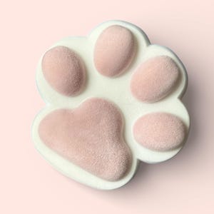 Puede incluir: Primer plano de un objeto con forma de pata de gato. La pata tiene un borde blanco y presenta cinco almohadillas rosas texturizadas. El fondo es de un rosa suave, creando una estética cohesiva y suave. El artículo es probablemente una pieza decorativa o un artículo funcional.