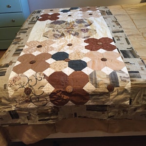 May include: A handmade quilt featuring a floral pattern in shades of brown, beige, and cream. The quilt is adorned with large, wooden buttons at the center of each flower. The quilt is placed on a bed with a patterned bedspread.