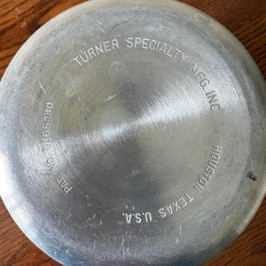 May include: Close-up of the bottom of a metal pan with the text "TURNER SPECIALTY MFG INC HOUSTON TEXAS U.S.A." The pan has a circular brushed metal design with the text "PAT NO. D166480".