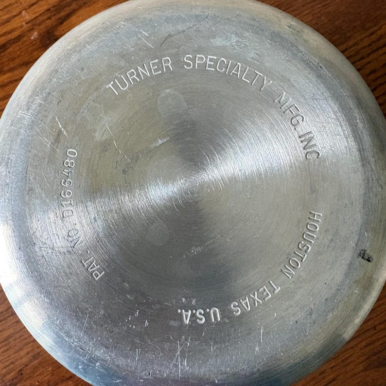 May include: Close-up of the bottom of a metal pan with the text "TURNER SPECIALTY MFG INC HOUSTON TEXAS U.S.A." The pan has a circular brushed metal design with the text "PAT NO. D166480".