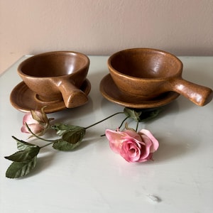 May include: Two brown ceramic soup bowls with handles, each resting on a matching saucer. The bowls have a speckled texture. A pink rose and green leaves are placed near the bowls.