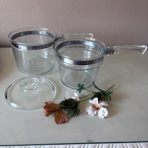 May include: Two clear glass cooking pots with silver-toned metal rims and transparent glass handles. One pot features a side handle, while the other is a deep bowl. A matching clear glass lid is included, accompanied by artificial flowers.