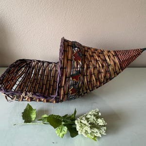 May include: A woven cornucopia basket in shades of brown, purple, and yellow, decorated with leaf accents. A green and white floral stem lies below the basket. The basket is designed to hold items.