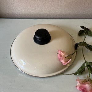 May include: Cream-colored, round pot lid with a black knob. The lid has a silver rim and is set on a light blue surface. A sprig of pink roses with green leaves is placed near the lid, adding a touch of color and contrast.