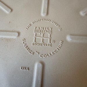 Puede incluir: Primer plano de la base de una fuente de horno de gres beige. El texto dice "The Pampered Chef Family Heritage Classics Stoneware Collection" y "USA". La fuente tiene una superficie texturizada y est&aacute; dise&ntilde;ada para cocinar.