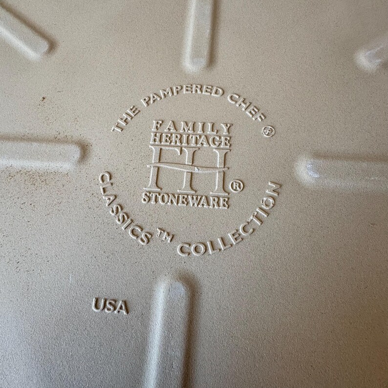 Puede incluir: Primer plano de la base de una fuente de horno de gres beige. El texto dice "The Pampered Chef Family Heritage Classics Stoneware Collection" y "USA". La fuente tiene una superficie texturizada y est&aacute; dise&ntilde;ada para cocinar.
