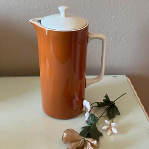 May include: A tall, orange ceramic pitcher with a white lid and handle. The pitcher has a glossy finish and a simple, elegant design. The handle is a light beige color. A sprig of flowers is in the foreground.
