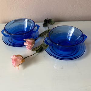 May include: Two cobalt blue glass soup bowls with matching saucers. Each bowl has two handles and sits on a round saucer. A pink rose and green leaves are placed between the bowls.