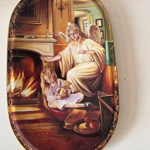 May include: Oval decorative plate featuring a serene scene of an angel and a child by a fireplace. The angel, in a flowing white gown, watches over the child, who holds a teddy bear. The plate has a gold trim and a warm color palette.