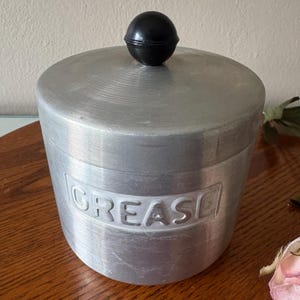 May include: A vintage aluminum grease container with a black knob on the lid. The word "GREASE" is embossed on the front of the container. The container is cylindrical and has a brushed metal finish.