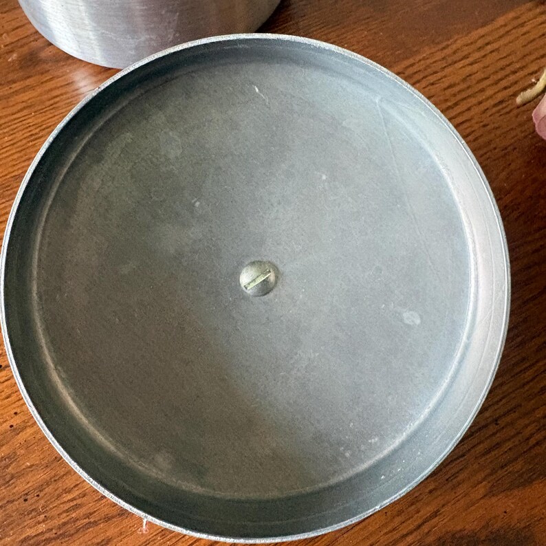 May include: A round, silver-colored metal lid with a screw in the center. The lid has a slightly textured surface and a raised edge. The lid is sitting on a wooden surface.