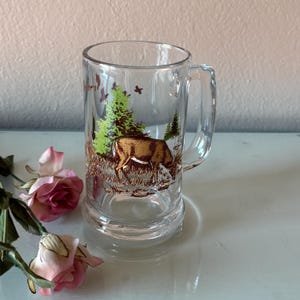 May include: Clear glass beer mug with a handle, featuring a deer graphic in a forest scene with green trees and brown grass. The mug has a wide base and a slightly flared top, perfect for enjoying a cold beverage.