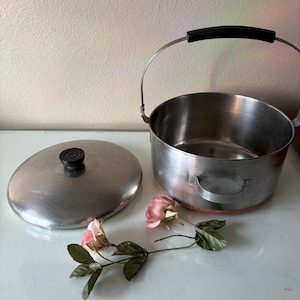 May include: A stainless steel cooking pot with a lid and a handle. The pot has a copper bottom and a black handle. A pink rose and green leaves are next to the pot and lid.