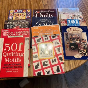 May include: A collection of quilting books and iron-on transfer leaflets. Titles include "505 Quilt Blocks", "American Patchwork & Quilting", "Quick Rotary Cutter Quilts", and "501 Quilting Motifs". The books feature colorful designs and patterns for quilting projects.