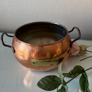May include: A copper potpourri bowl with two curved handles. The bowl has a rounded shape and a tarnished finish, with a small brass plate that reads "POTPOURRI". A pink rose and green leaves are visible in the background.