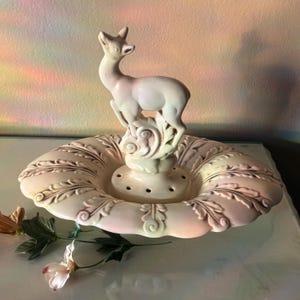 May include: A cream-colored ceramic centerpiece featuring a deer figurine atop a decorative base. The base has a scalloped edge with embossed leaf and scroll designs. The deer is light pink and stands on a pedestal with holes.