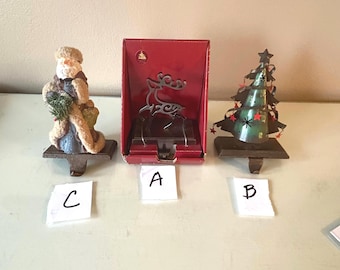May include: Three Christmas stocking holders: a Santa figurine on a brown base, a silver-toned metal reindeer in a red box labeled A, and a green metal tree with red star accents on a brown base labeled B. The Santa figurine is labeled C.