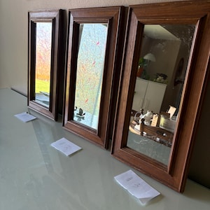May include: Three rectangular mirrors with dark brown wooden frames. The mirrors reflect the surrounding environment, including a glimpse of a yard and interior decor. Each mirror has a different design, with one featuring a floral pattern and another a decorative element at the bottom.