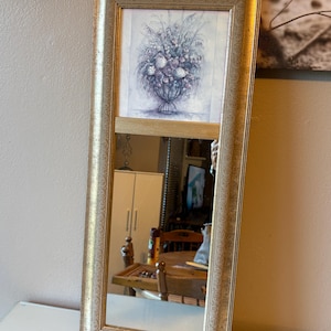 May include: A rectangular mirror with a gold-colored frame. The top section features a framed floral print in shades of blue and gray. The mirror reflects a room with furniture and other objects.