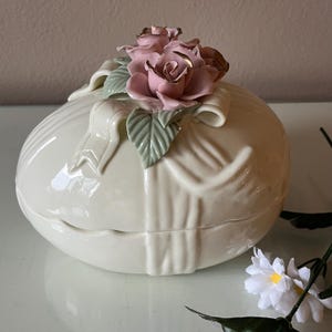 May include: A cream-colored ceramic trinket box with a lid, adorned with a cluster of pink roses and green leaves. The box has a ribbon design and a glossy finish, perfect for storing small items.