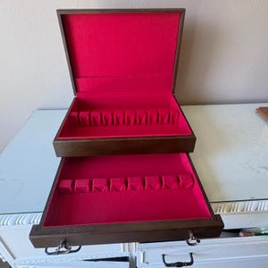 May include: A dark brown wooden flatware storage chest with a hinged lid and a pull-out drawer. The interior of both the upper compartment and the lower drawer is lined with bright red felt and features molded slots for organizing silverware.