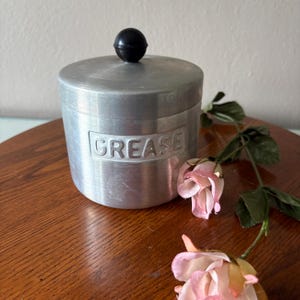 May include: A vintage aluminum grease container with a black knob on the lid. The word "GREASE" is embossed on the front. The container is cylindrical and sits on a wooden surface, with pink flowers in the foreground.