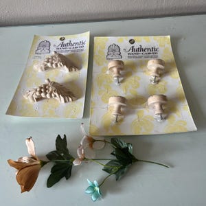 May include: Two packages of "Authentic Hand-Carved" wood appliques, one with grape cluster designs and the other with four knobs. A stem of artificial flowers with orange, white, and blue blossoms is also present.