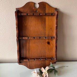 May include: A vintage wooden spoon rack with three shelves. The rack is made of dark stained wood and has a decorative top with a hole. The edges of the rack have a rustic, uneven appearance. The shelves have small slots to hold spoons.