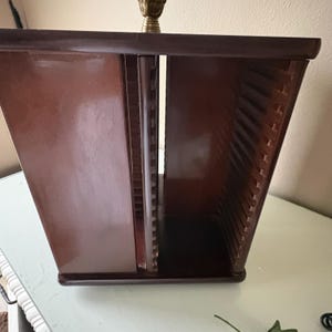 May include: A dark brown wooden media storage rack with a glossy finish, featuring a central vertical divider and multiple horizontal slots on both sides for organizing discs. A small metallic gold-toned finial is visible at the very top center.