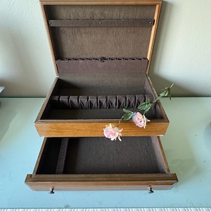 May include: A wooden jewelry box with a hinged lid and a pull-out drawer. The interior is lined with dark brown fabric, featuring a ring holder slot and a necklace bar. A decorative artificial pink rose stem is placed across the open compartments.