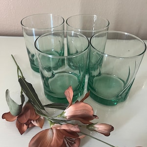 May include: Four clear, green-tinted drinking glasses with a rounded base and straight sides. The glasses are arranged on a white surface, with a sprig of brown and pink flowers in the foreground. The glasses are suitable for water, juice, or cocktails.