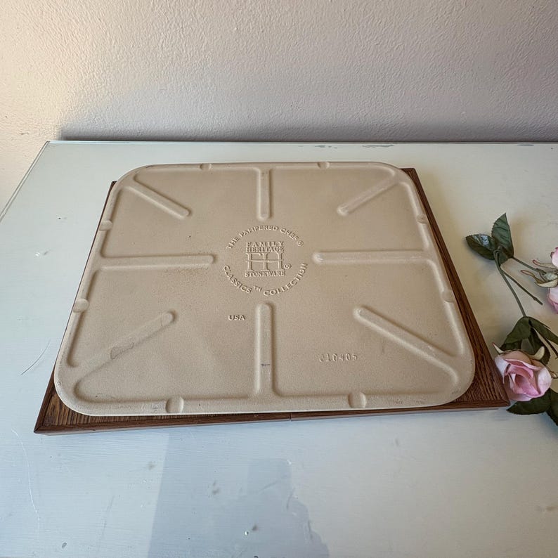 Puede incluir: Una piedra para hornear cuadrada de gres blanquecino con una base de madera marr&oacute;n. La piedra tiene una superficie texturizada y se ve el texto "THE PAMPERED CHEF" y "FAMILY HERITAGE STONEWARE CLASSICS COLLECTION USA". Unas rosas rosas est&aacute;n en el fondo.