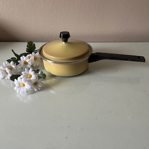 May include: A vintage yellow saucepan with a black handle and lid. The lid has a black knob. The saucepan is sitting next to a bouquet of white daisies with yellow centers and green leaves.
