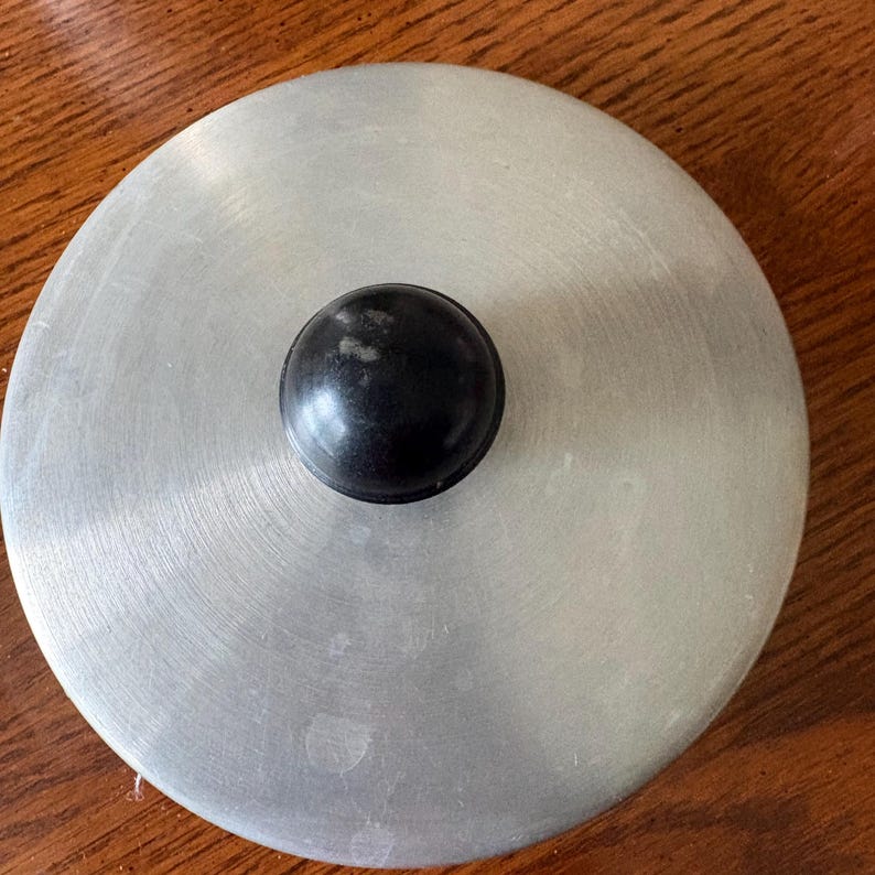May include: A circular, brushed metal lid with a black, rounded knob in the center. The lid has a series of concentric circular patterns radiating from the center. The knob is a solid, dark color.