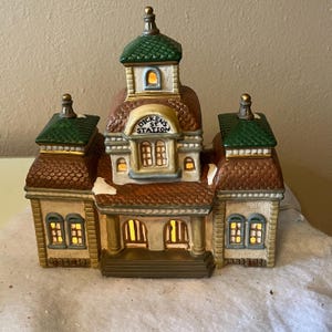 May include: A ceramic Christmas village building with the text "Dickens St. Station". The building has a brown roof, green accents, and lit windows. The building is beige with brown and green details.