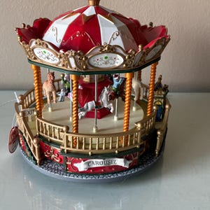 May include: A miniature musical carousel featuring a red and white striped roof, gold spiral pillars, and a platform with various animal figures. The base is decorated with a banner labeled CAROUSEL and an EXIT sign, with a tag reading I'M MUSICAL.
