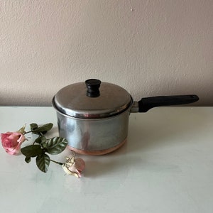 May include: A stainless steel saucepan with a copper bottom, black handle, and a lid with a black knob. The pan is on a white surface with pink roses.