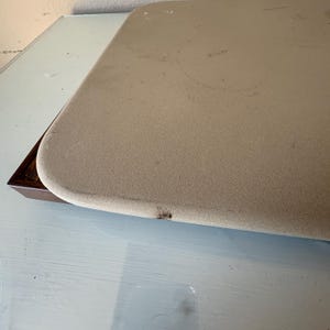Puede incluir: Primer plano de una mesa beige rectangular con esquinas redondeadas. La mesa tiene una superficie texturizada y est&aacute; sobre una base de madera marr&oacute;n oscuro. La mesa est&aacute; sobre una superficie azul claro.