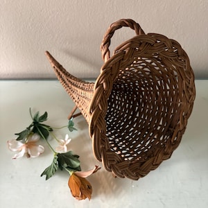 May include: A brown woven wicker cornucopia basket featuring a wide, circular opening with a braided rim and a tapered, curved tail. The basket is positioned next to a small, artificial floral stem with white and orange petals and green leaves.