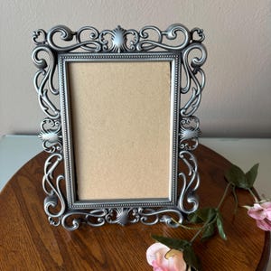 May include: A silver-colored rectangular picture frame with ornate scrollwork and shell-like accents. The frame has a neutral-colored backing and is displayed on a wooden surface. Pink flowers are in the foreground.