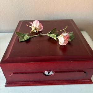 May include: A rectangular, dark red wooden jewelry box with a keyhole lock. Two pink roses with green leaves rest on the box's top. The box has a decorative, recessed panel on the front.