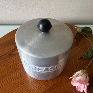 May include: A vintage aluminum grease container with a black knob on the lid. The word "GREASE" is embossed on the side. The container sits on a wooden surface with a pink rose.