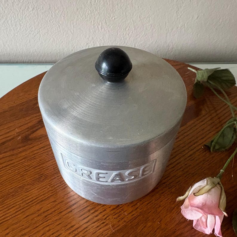 May include: A vintage aluminum grease container with a black knob on the lid. The word "GREASE" is embossed on the side. The container sits on a wooden surface with a pink rose.